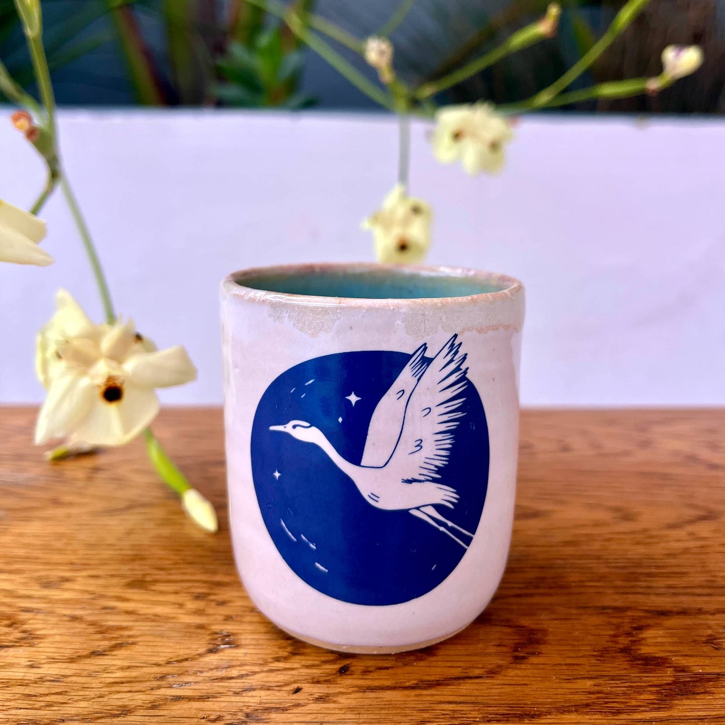 Ceramic cup with a blue design of a bird on a wooden surface with flowers in the background