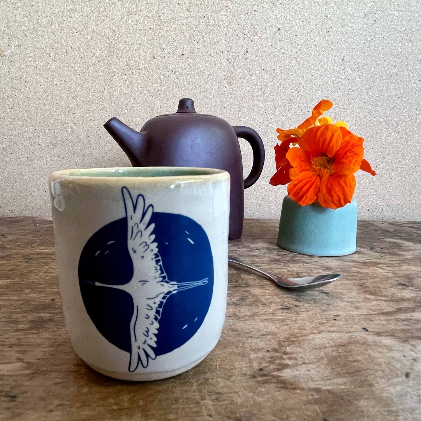 Ceramic mug with blue design, teapot, spoon, and orange flowers on a wooden surface.