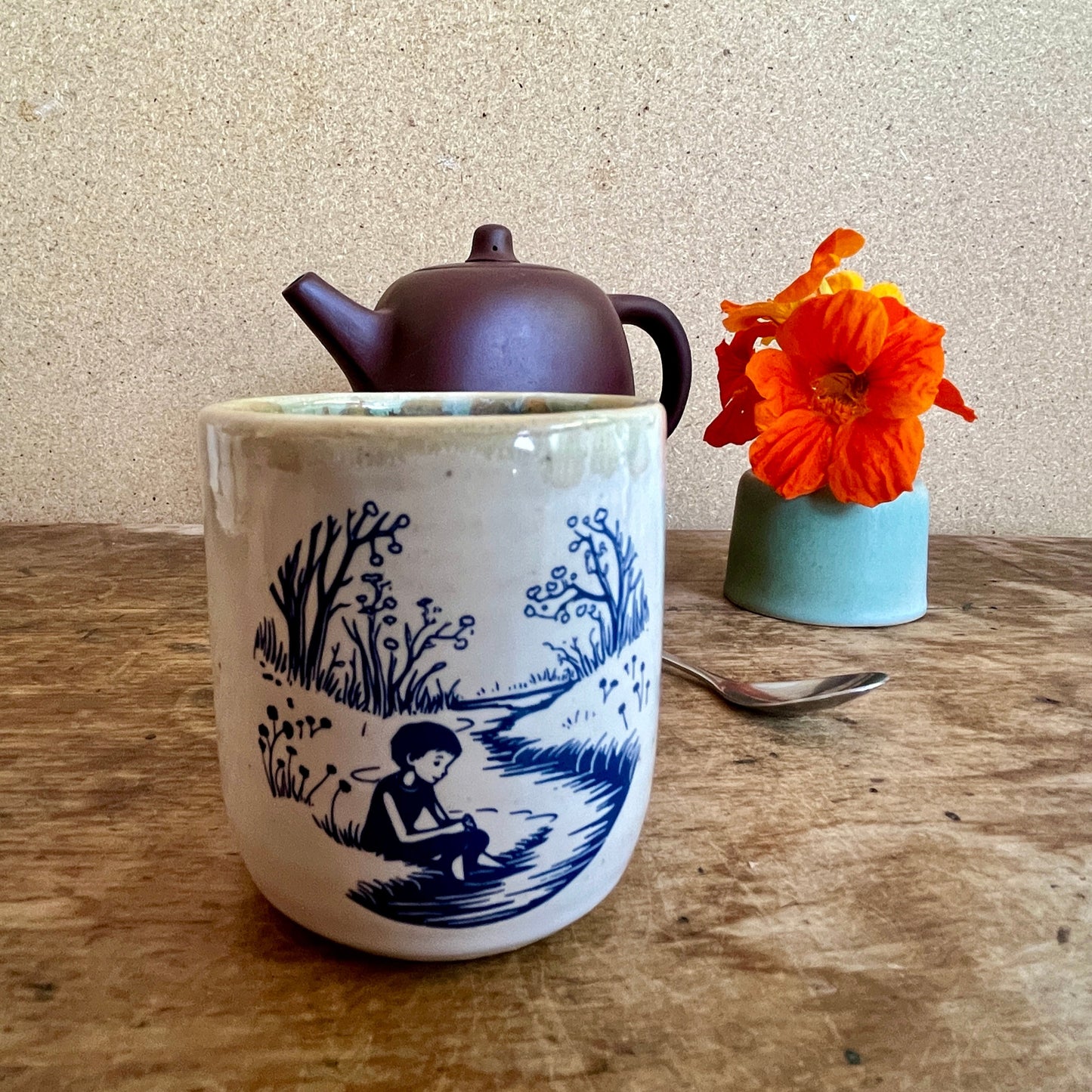 Ceramic mug with blue design, teapot, and orange flower on a wooden table.