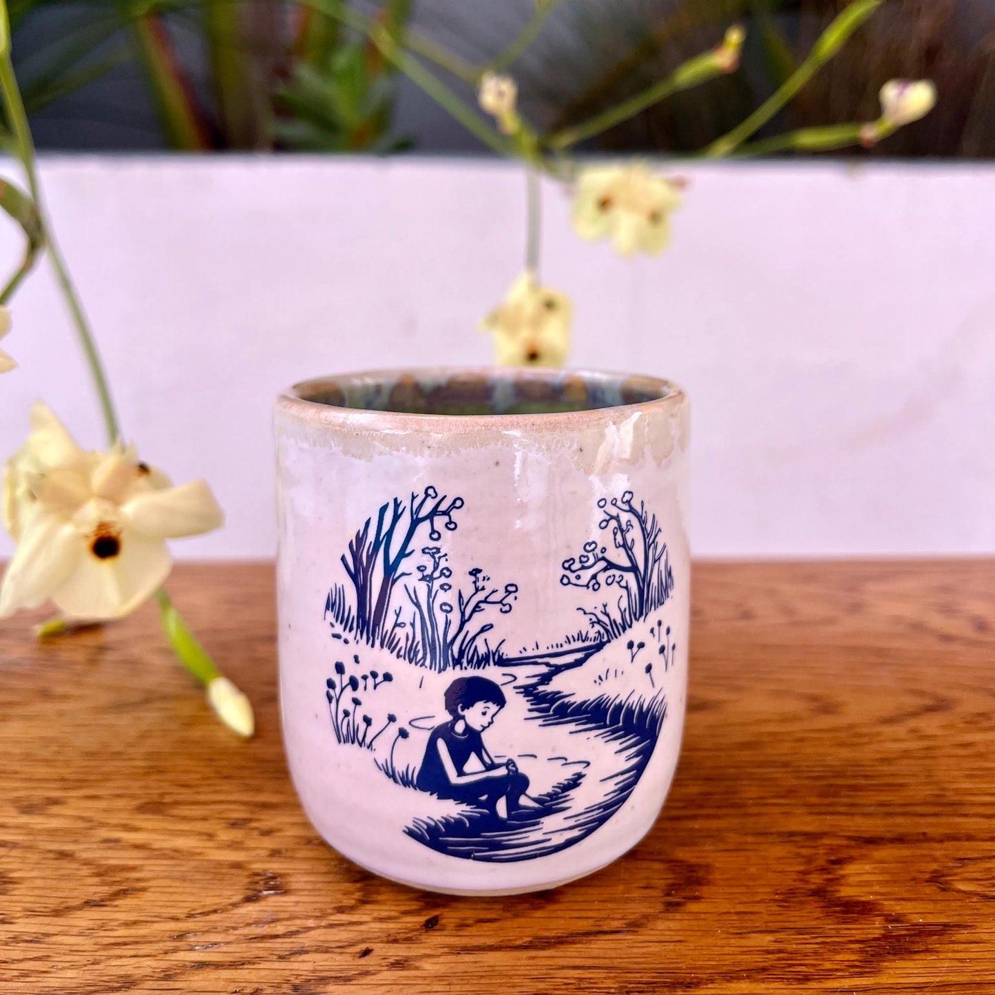 Ceramic mug with blue landscape and figure design on a wooden surface with flowers in the background