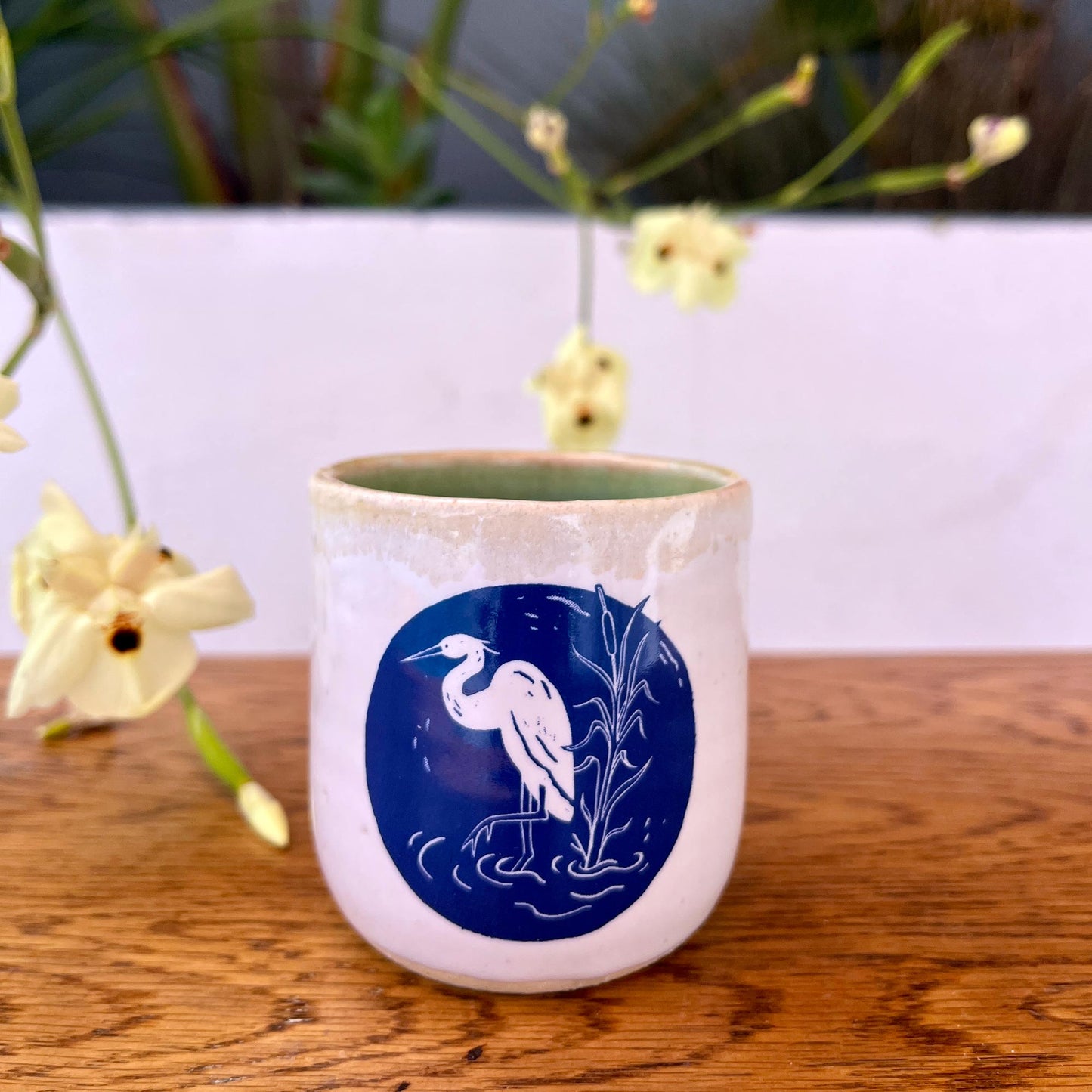 Ceramic mug with a blue design of a heron on a wooden surface with flowers in the background