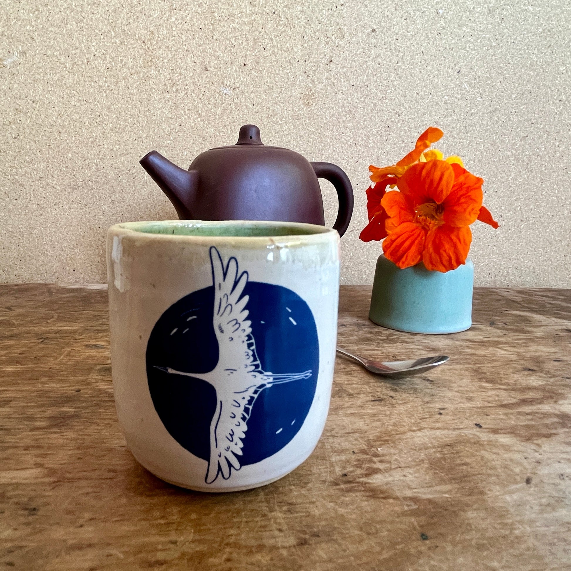 Ceramic mug with bird design, teapot, and flowers on a wooden table.