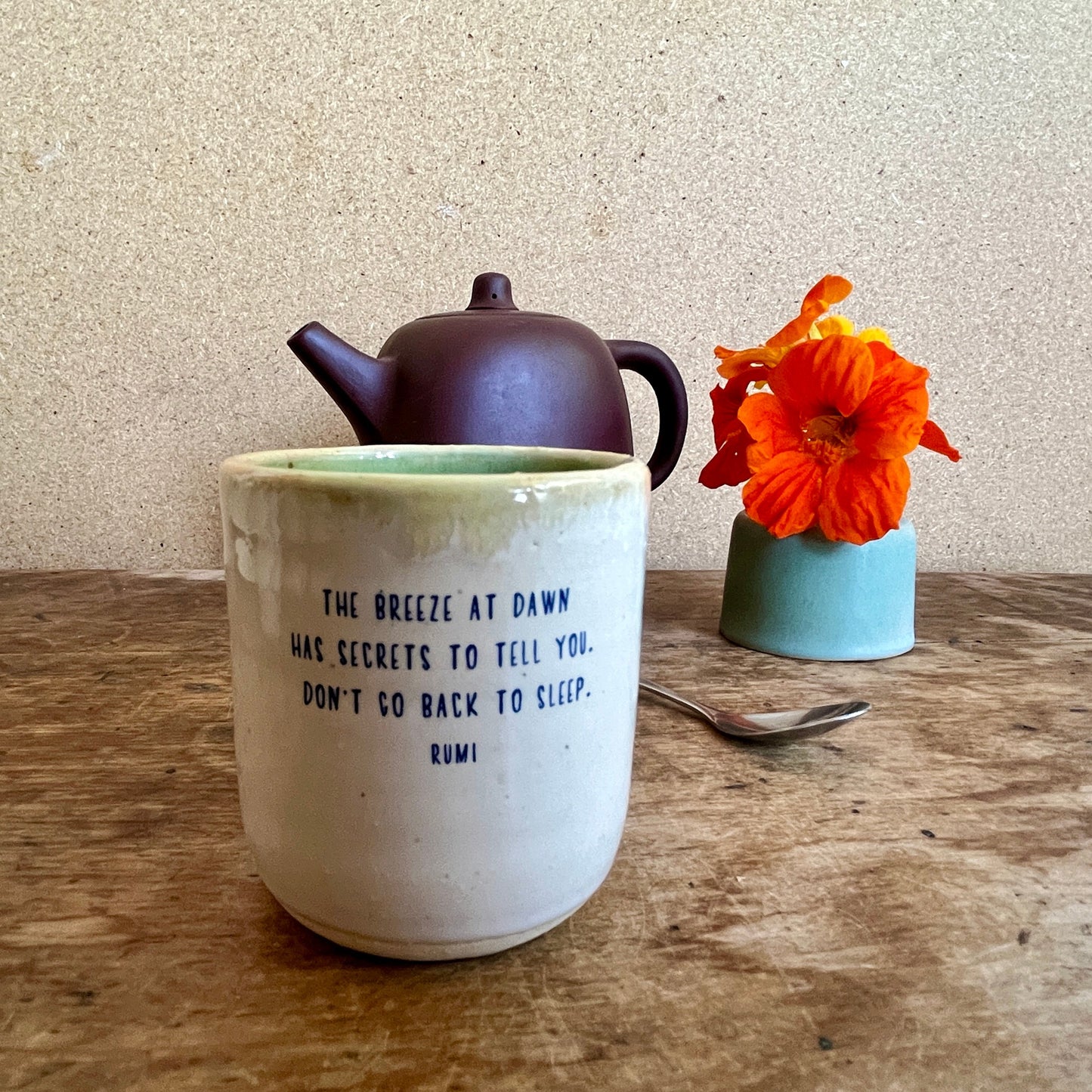 Ceramic mug with text, teapot, and orange flower on a wooden surface